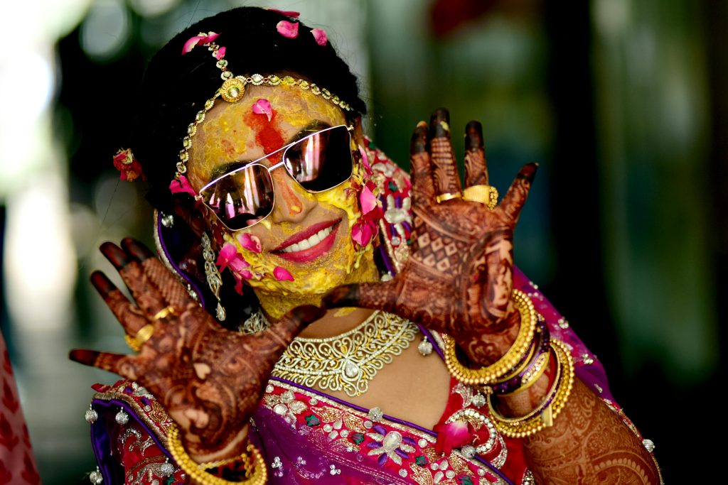 A joyful Indian bride adorned with traditional jewelry and mehndi, smiling in sunglasses.