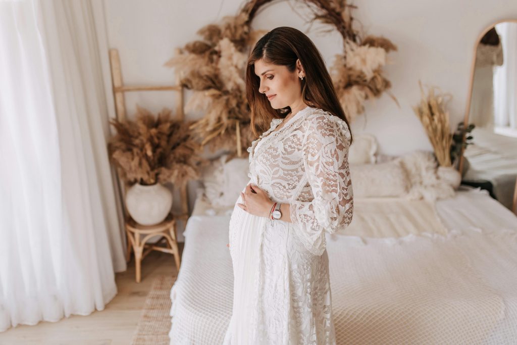 A pregnant woman in a white lace dress stands in a bohemian-style bedroom, embracing maternity.